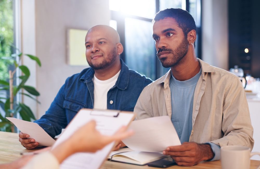 Two men in casual attire sit at a table, holding documents, during a meeting in a bright, modern office space.