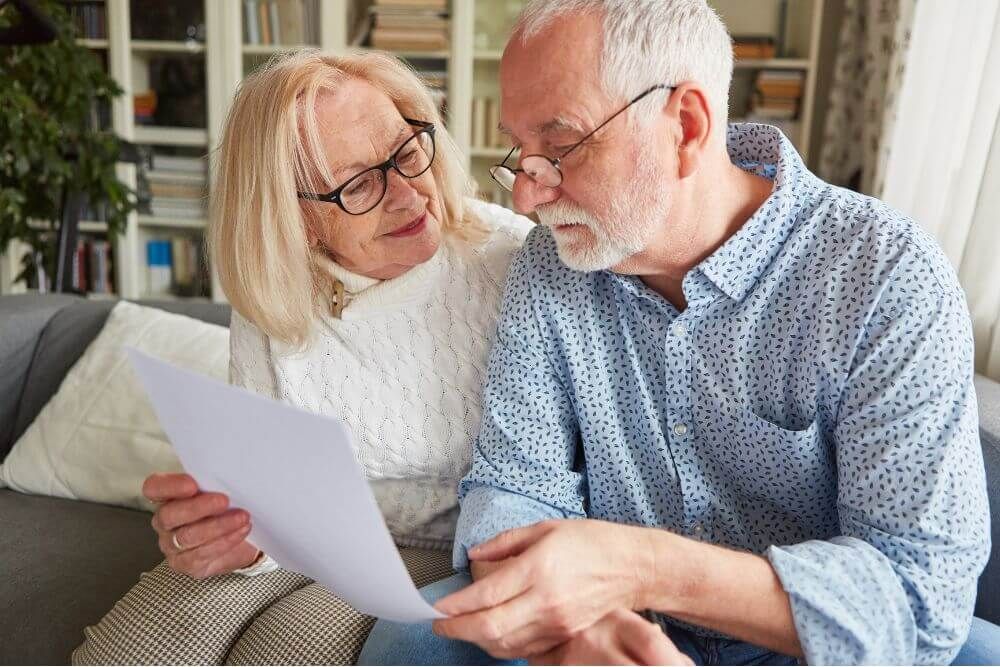 An elderly couple sits closely together on a couch, examining a piece of paper. The woman smiles at the man, who is focused on the document, in a cozy, well-lit living room.