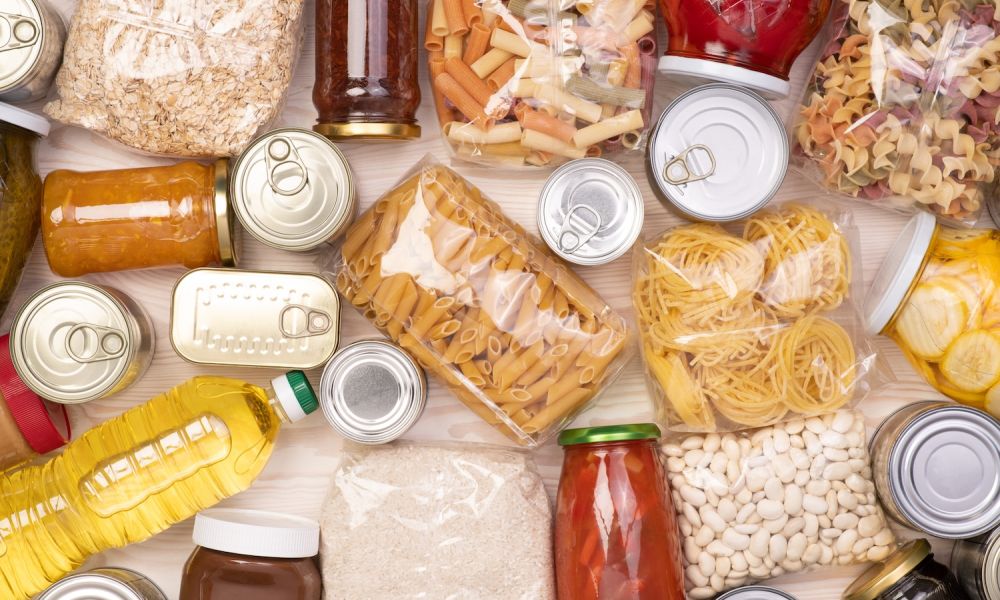 Assorted pantry staples including pasta, canned goods, and bottled items arranged on a wooden surface.