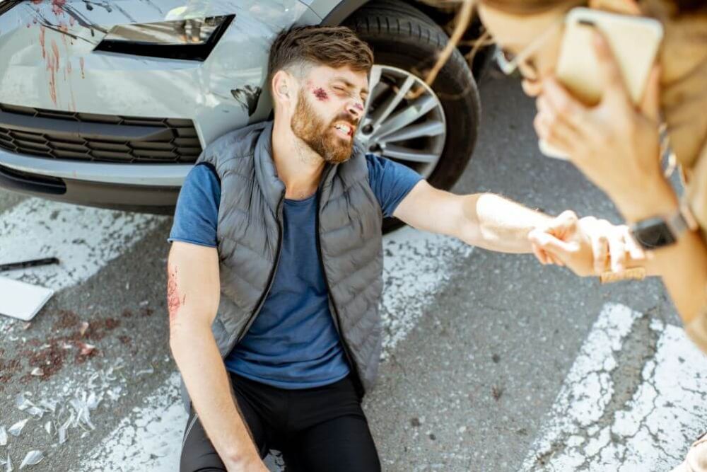 A man with facial injuries sits on the ground, leaning against a car with a damaged front bumper. Glass shards are scattered nearby. Another person uses a phone for assistance.