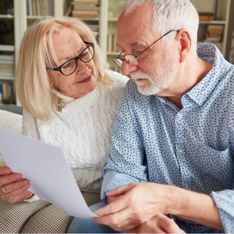 An elderly couple sits closely together on a couch, examining a piece of paper. The woman smiles at the man, who is focused on the document, in a cozy, well-lit living room.