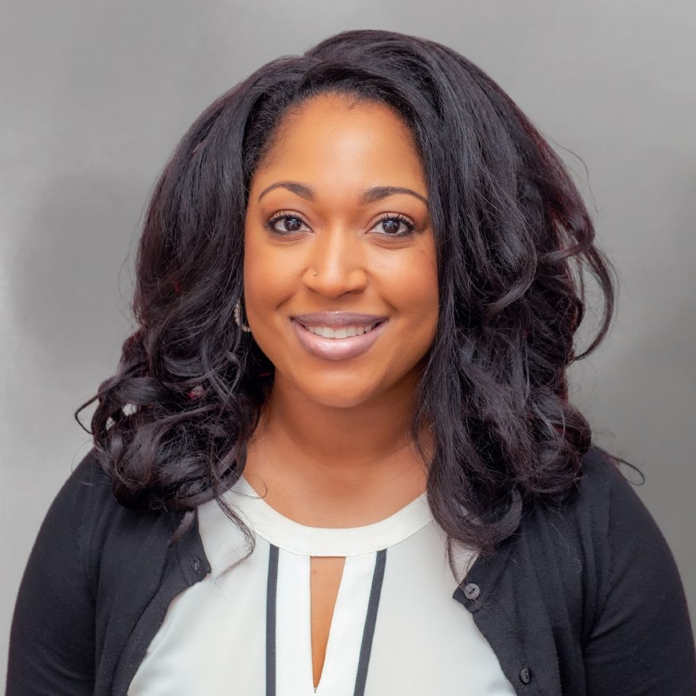 Willonda Ashton smiling in professional attire against a gray background, with long, wavy hair.