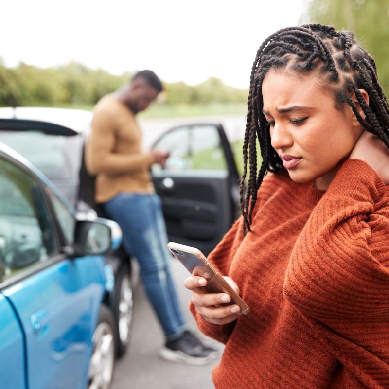 Woman on phone after car collision, rubbing her neck; man in background checking phone near car door.