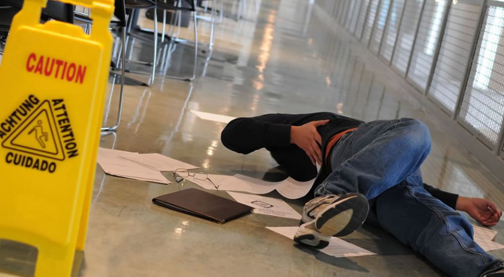 Person slipped on wet floor near caution sign in hallway, surrounded by scattered papers and belongings.