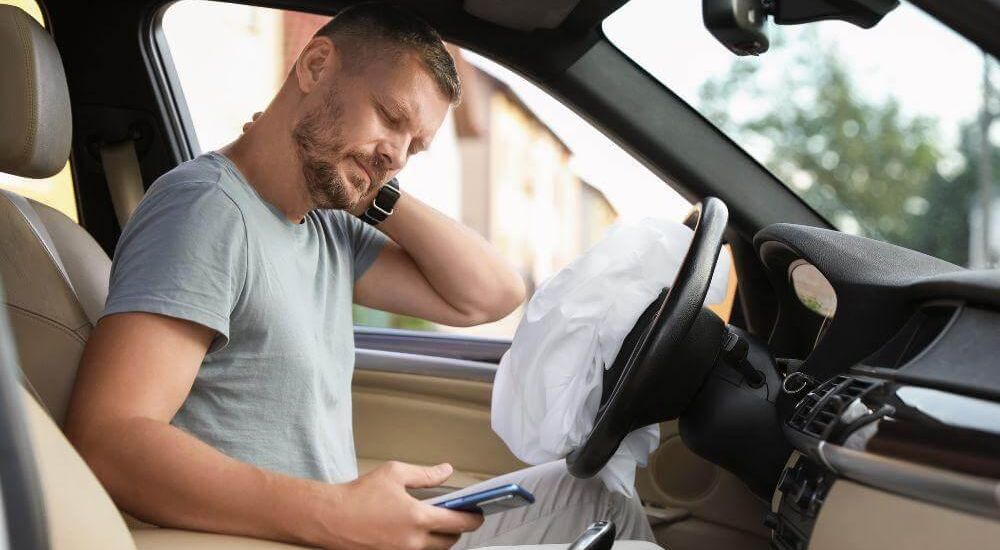 Man seated in a car with the front airbag deployed, looking down at his smartphone.