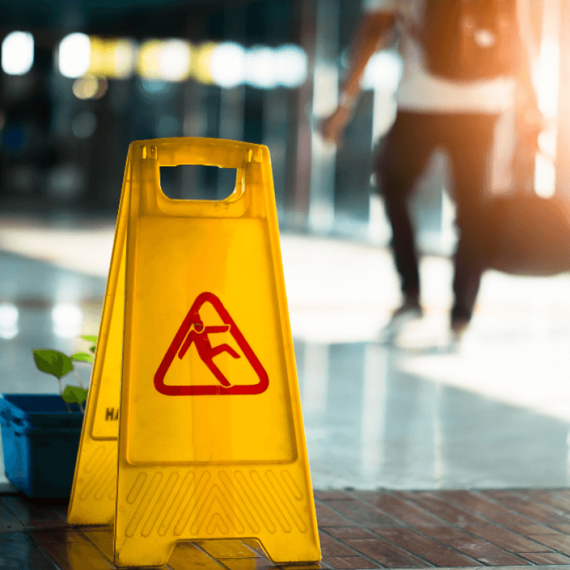 A yellow caution sign indicating a slip hazard stands on a polished floor, while a person with a bag walks past in a well-lit indoor area. A small plant is nearby.