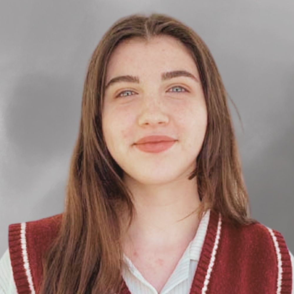 Megan Rathmell with long brown hair in a maroon sweater vest, smiling against a gray background.