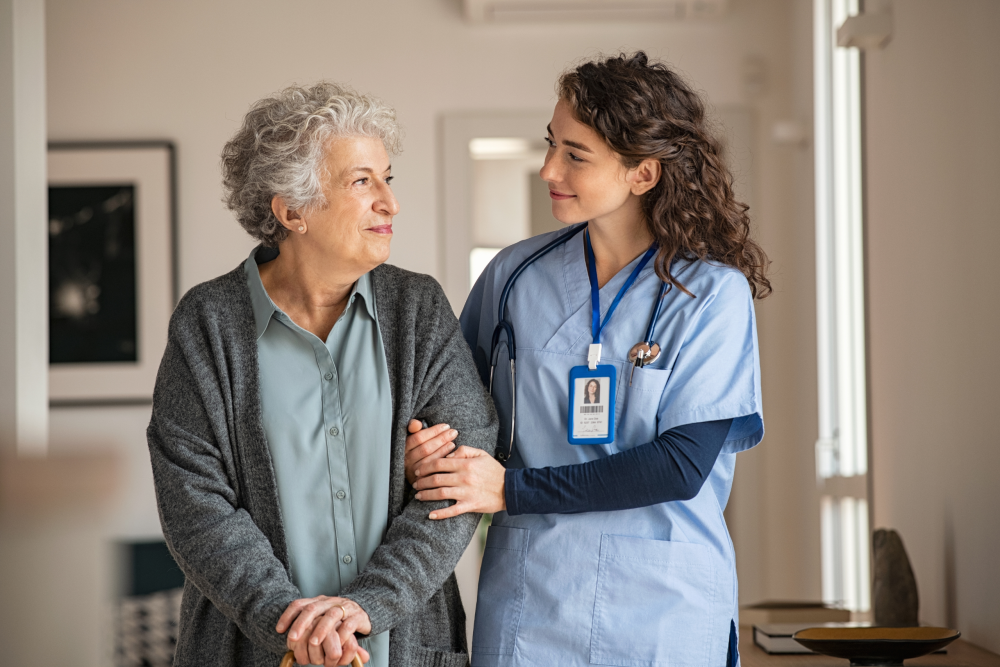 An elderly woman with curly gray hair, wearing a cardigan over a blouse, is standing and smiling while looking to her side. Beside her, a compassionate female nurse in a light blue scrub and with a badge around her neck is standing close, providing support with a gentle hand on the woman's arm. They appear to be in a well-lit nursing home environment with minimal decor.