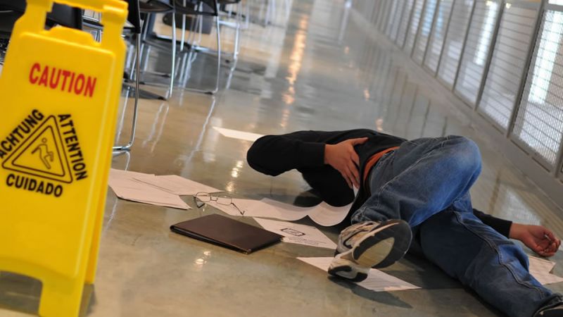 Person slipped on wet floor near caution sign in hallway, surrounded by scattered papers and belongings.