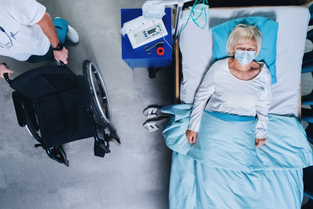 Overhead view of masked doctor arriving with wheelchair next to elderly nursing home resident lying in bed