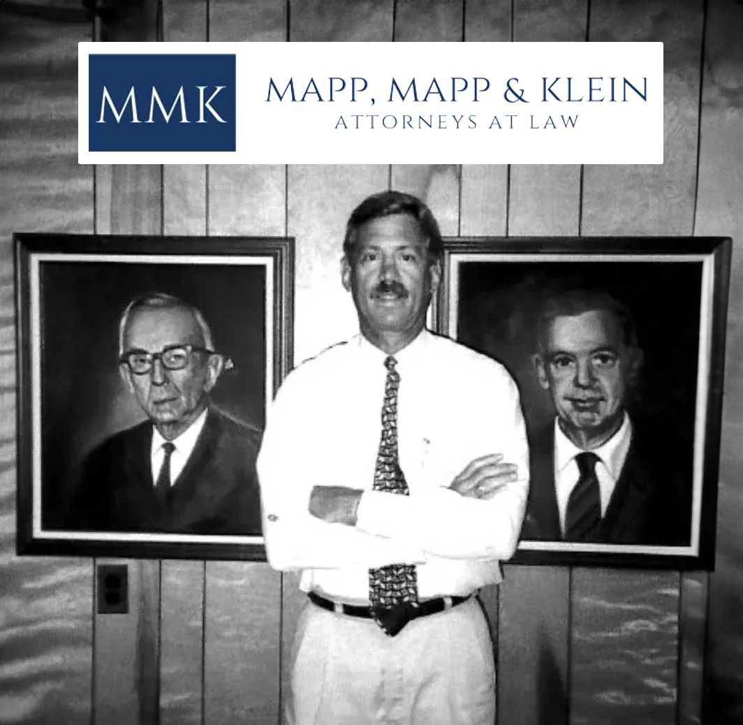 Hombre de pie entre dos retratos en una oficina de abogados, con el logotipo de Mapp, Mapp & Klein encima.