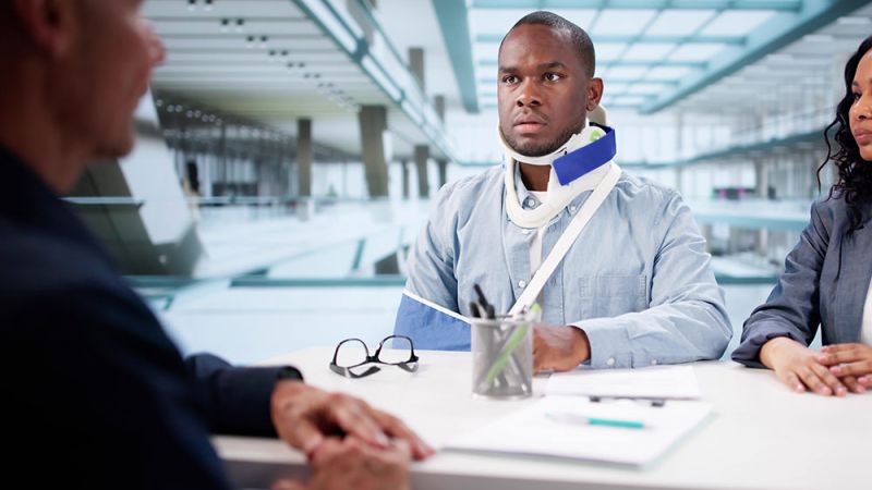 Man with neck brace and arm sling discussing with professionals in office setting.