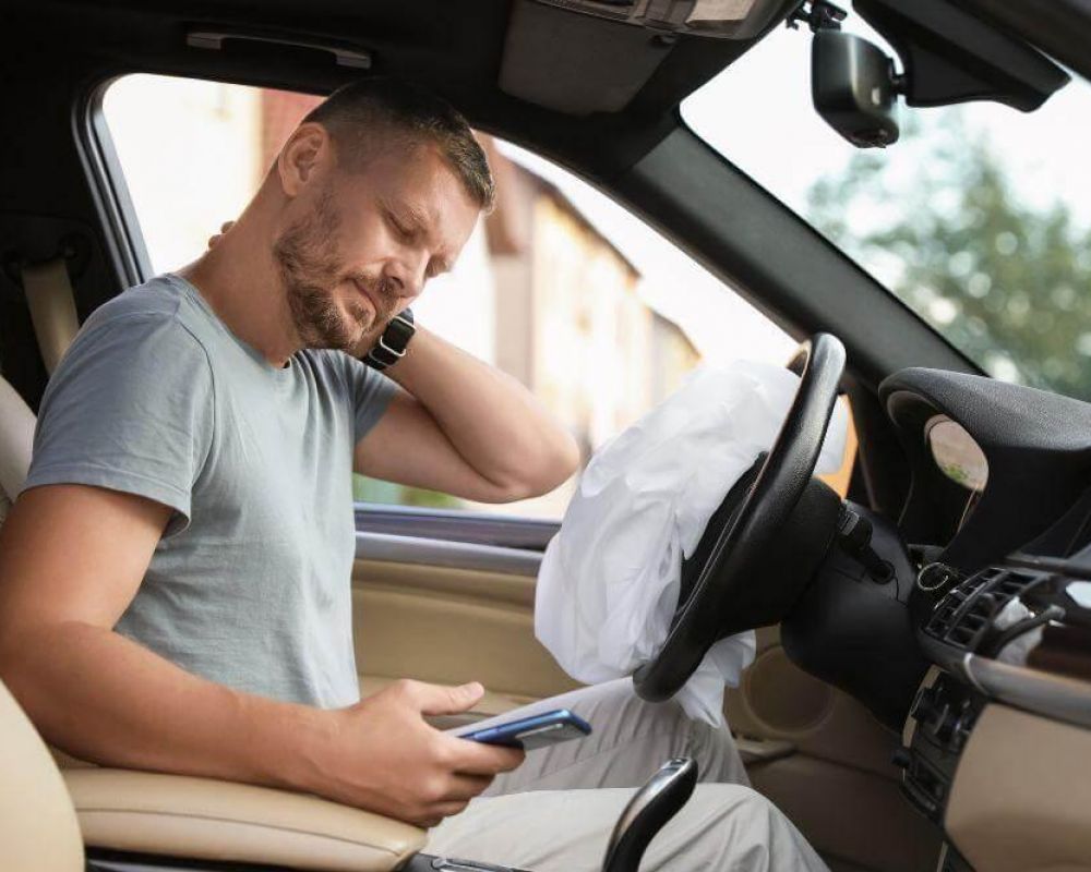 Man seated in a car with the front airbag deployed, looking down at his smartphone.