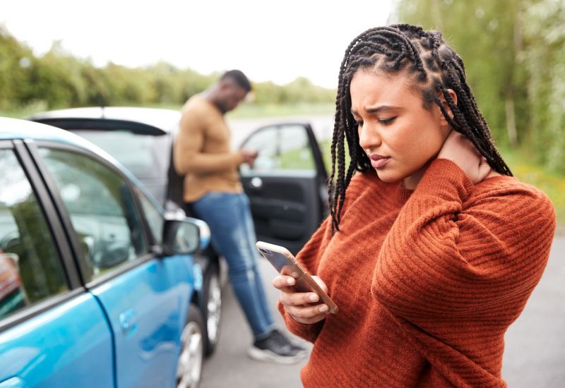 Woman on phone after car collision, rubbing her neck; man in background checking phone near car door.