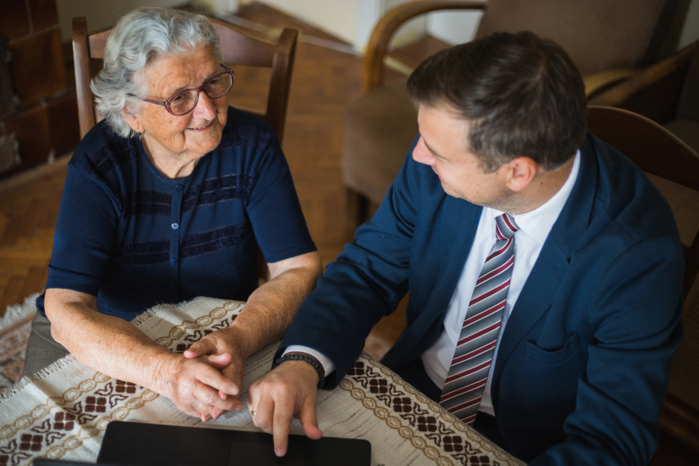Elderly woman smiling and holding hands with a man in a suit, as he points to a document on a tablet during a legal consultation.