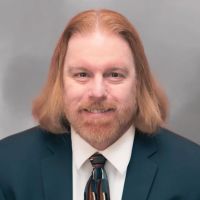 Jonathan R. DeLoatche with long hair and beard wearing a dark suit and colorful tie, smiling against a neutral background. Business portrait.