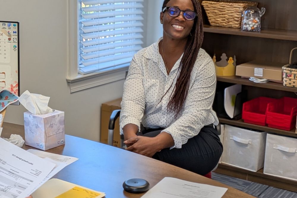 Smiling woman in a polka dot shirt sitting in an office, surrounded by paperwork and office supplies.