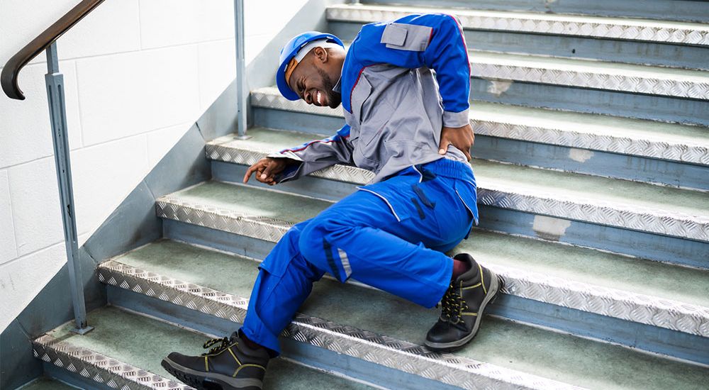 Worker in overalls and helmet falls on stairs, clutching back in pain. Workplace safety accident concept.