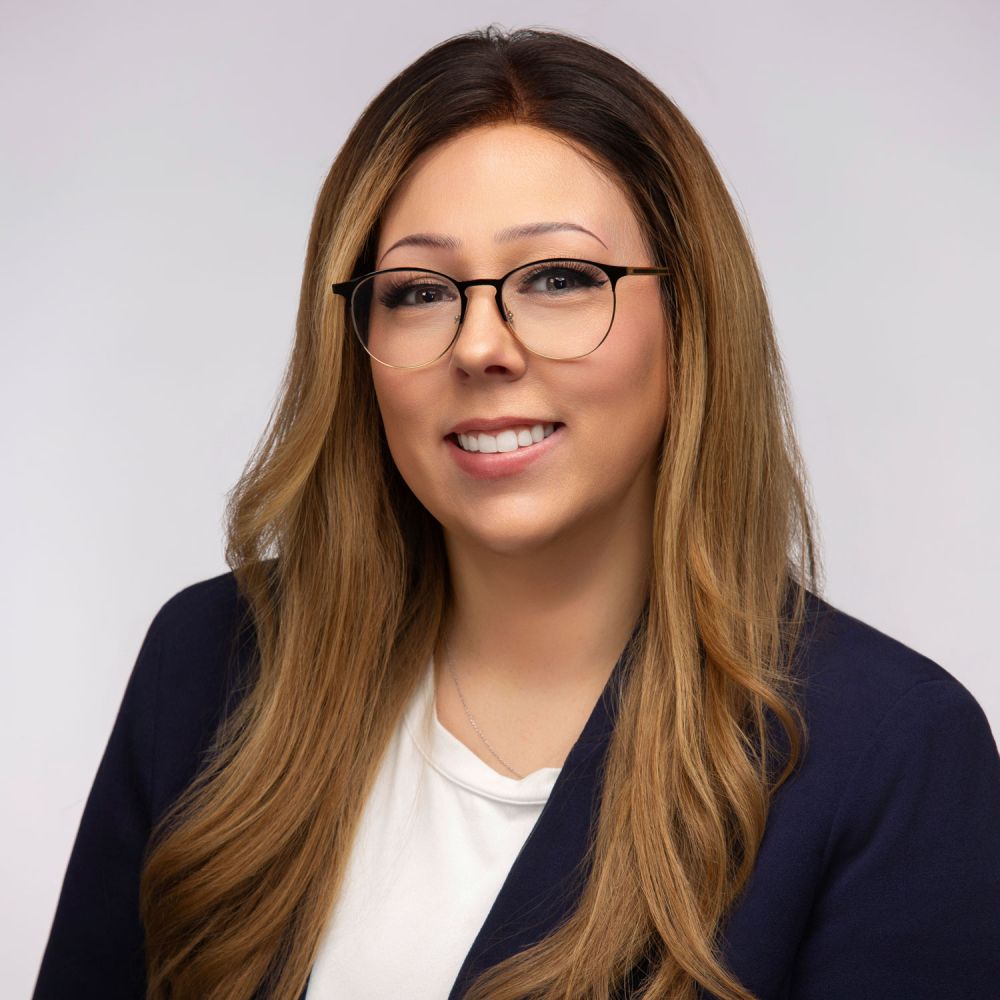 Jamie Imparato with glasses and long hair, wearing a navy blazer and white top, neutral background.