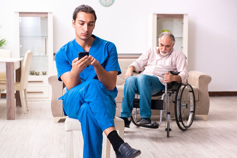 Caregiver in blue scrubs on phone while an elderly man in a wheelchair observes, in a home setting.