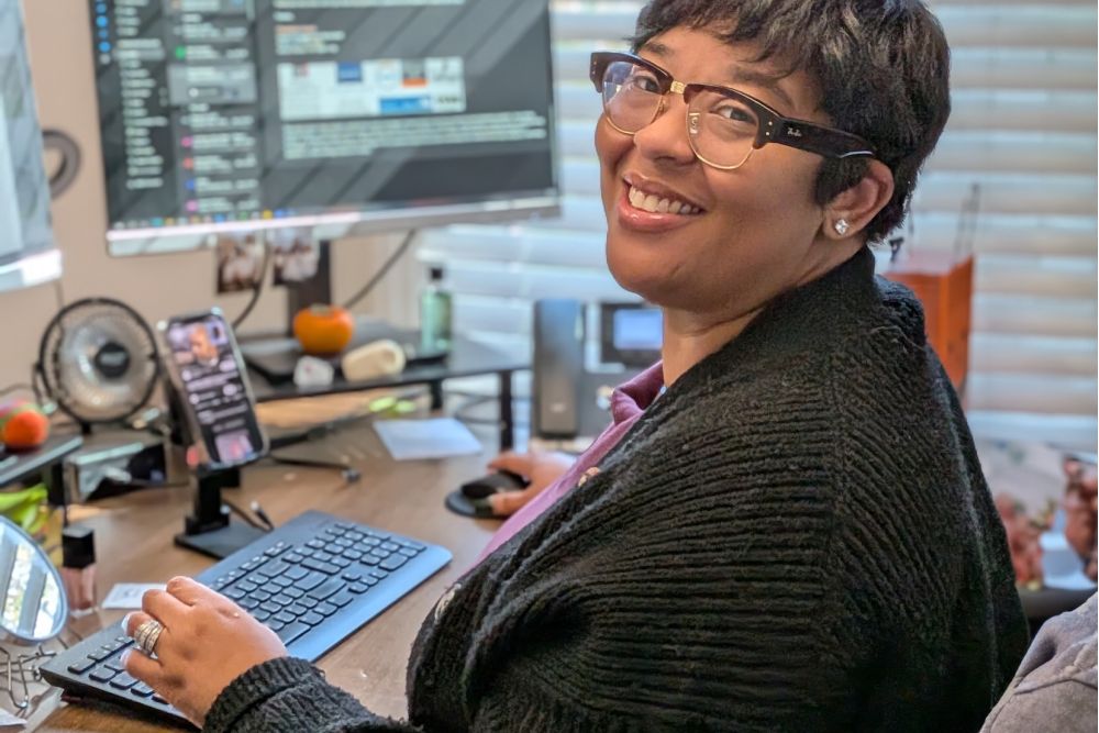 Smiling person using a computer in a home office with multiple screens and a cozy setup.