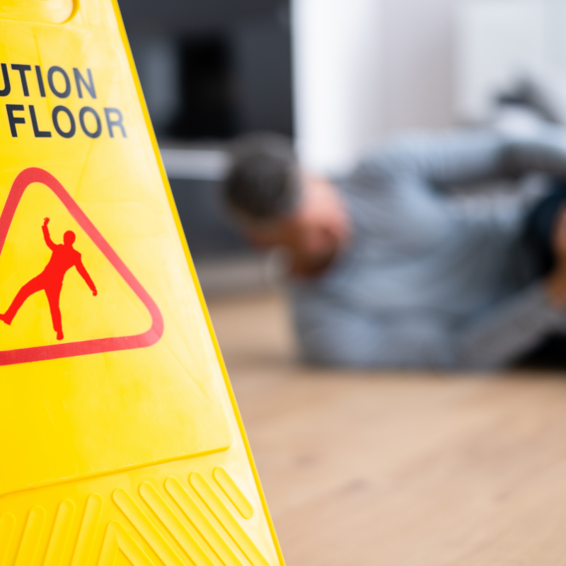 Foreground focus on a 'Caution Wet Floor' sign with a blurry background of a man holding his knee on the floor.