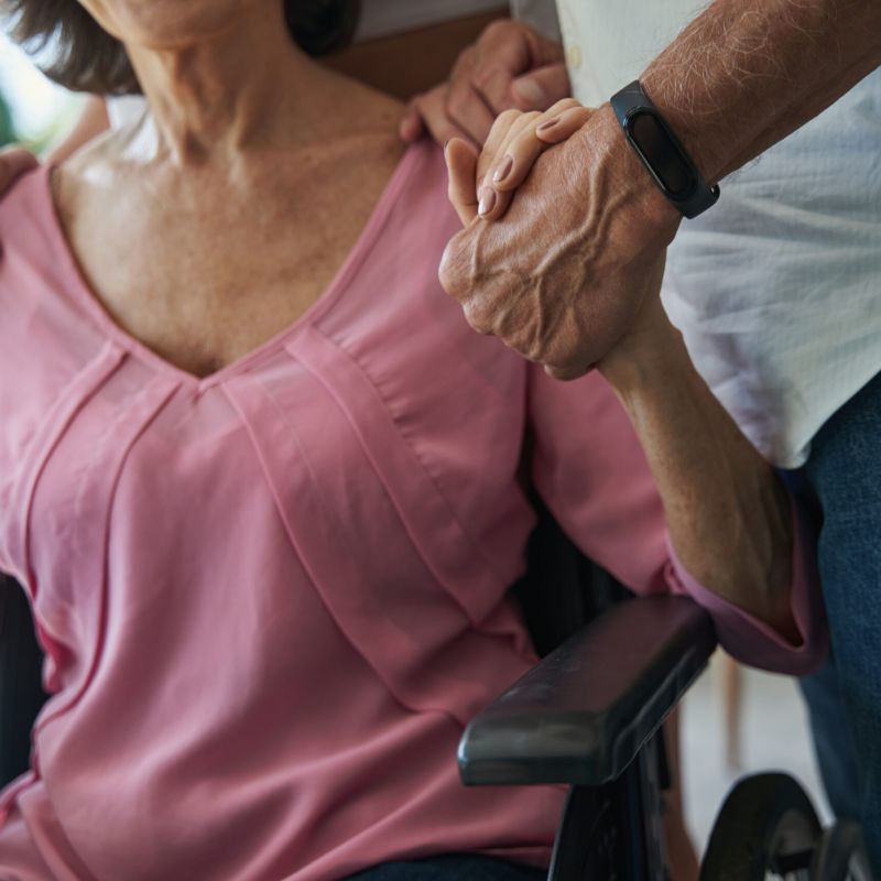 Close-up of a caregiver holding an elderly woman's hand while she sits in a wheelchair, emphasizing the importance of movement in nursing home care.