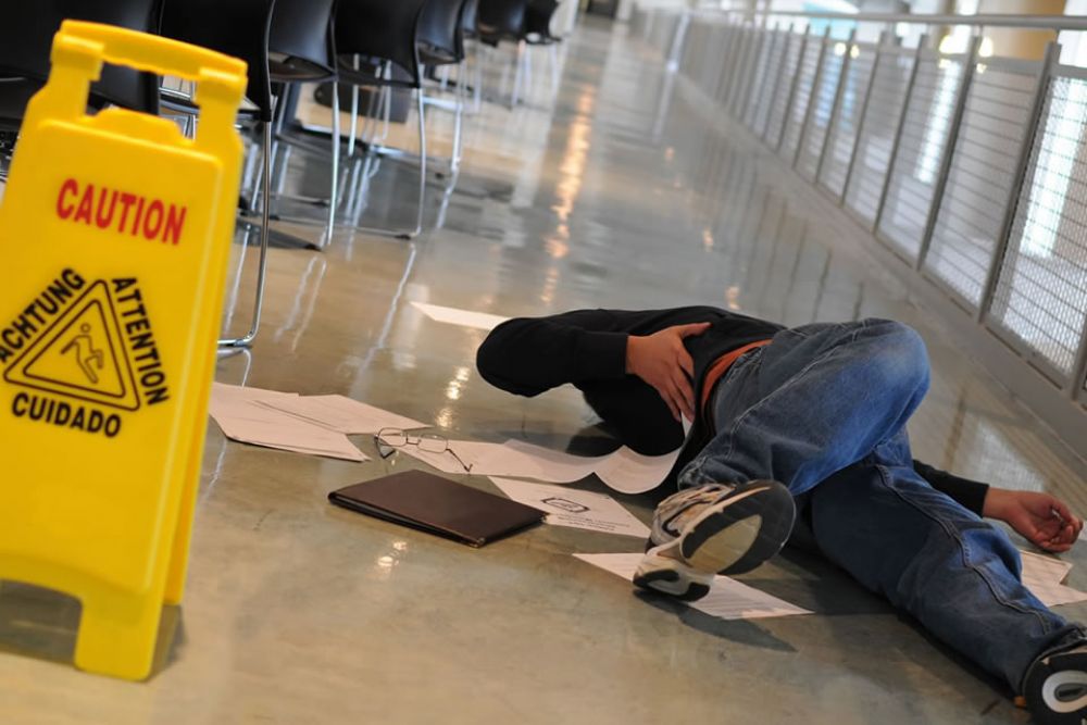 Person slipped on wet floor near caution sign in hallway, surrounded by scattered papers and belongings.