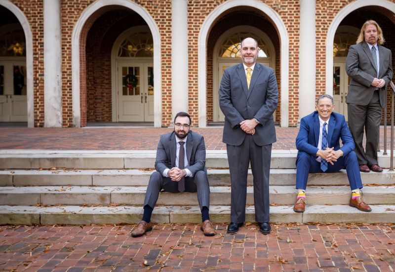 Four men in suits pose in front of a brick building with arches, two sitting and two standing on steps.
