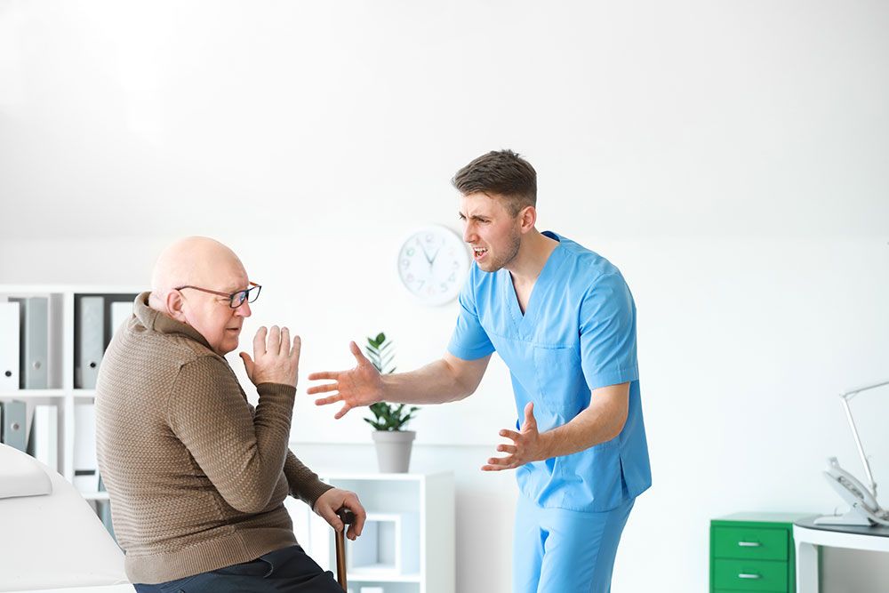 Healthcare professional in blue scrubs communicating with an elderly patient in a doctor's office setting.
