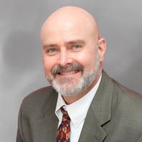 Robert Frazier with a beard in a suit smiling at the camera against a gray background.
