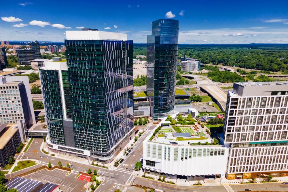 Aerial view of modern glass skyscrapers and lush greenery in an urban landscape under a clear blue sky.