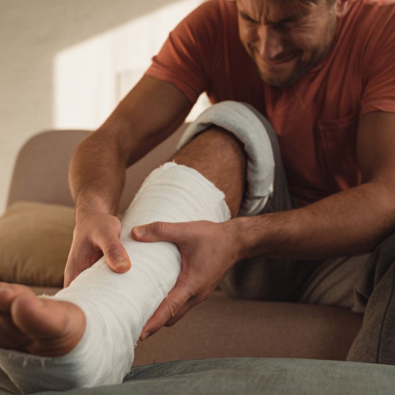 Man sitting on a couch holding his bandaged leg in pain, representing the challenges of recovering from an injury and navigating claims.
