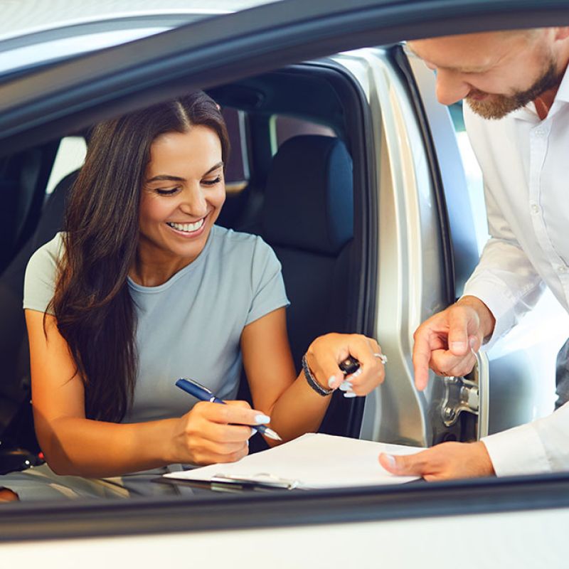 Woman signing documents in car with salesperson assisting.
