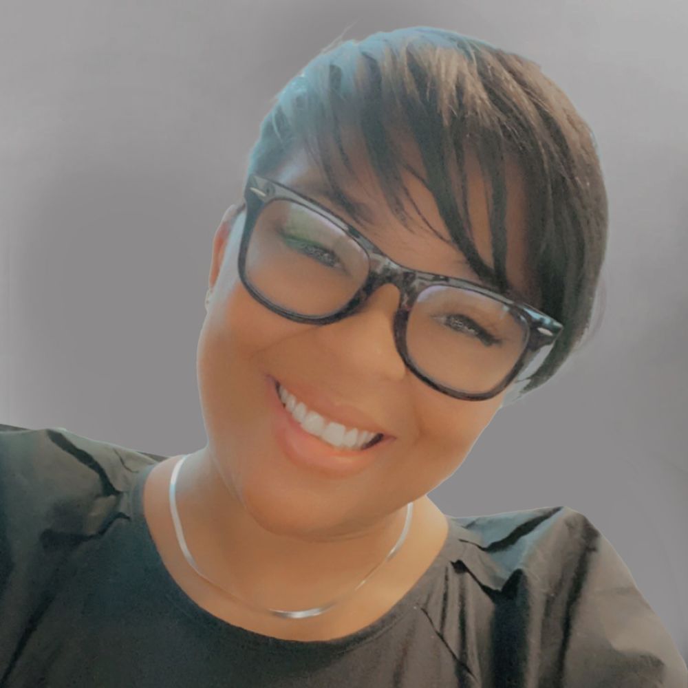 Smiling woman with short hair and glasses, wearing a black top and necklace, against a gray background.