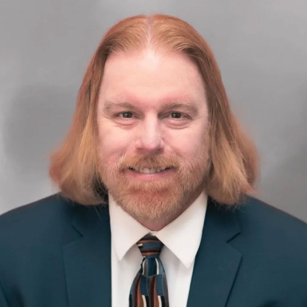 Jonathan R. DeLoatche with long hair and beard wearing a dark suit and colorful tie, smiling against a neutral background. Business portrait.