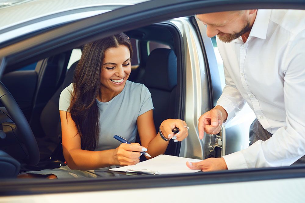 Woman signing documents in car with salesperson assisting.