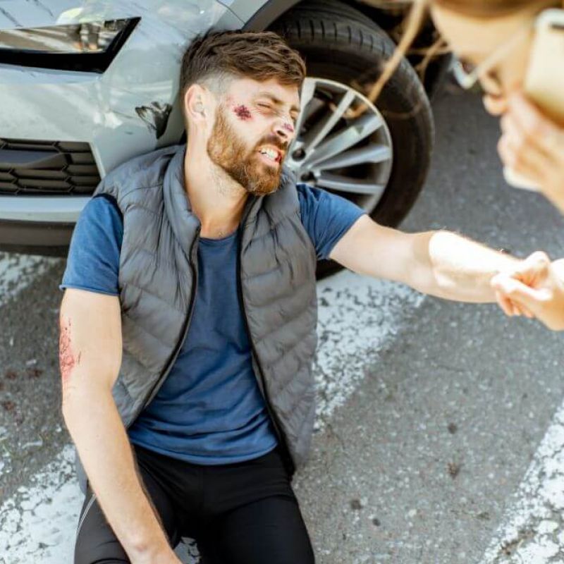A man with facial injuries sits on the ground, leaning against a car with a damaged front bumper. Glass shards are scattered nearby. Another person uses a phone for assistance.