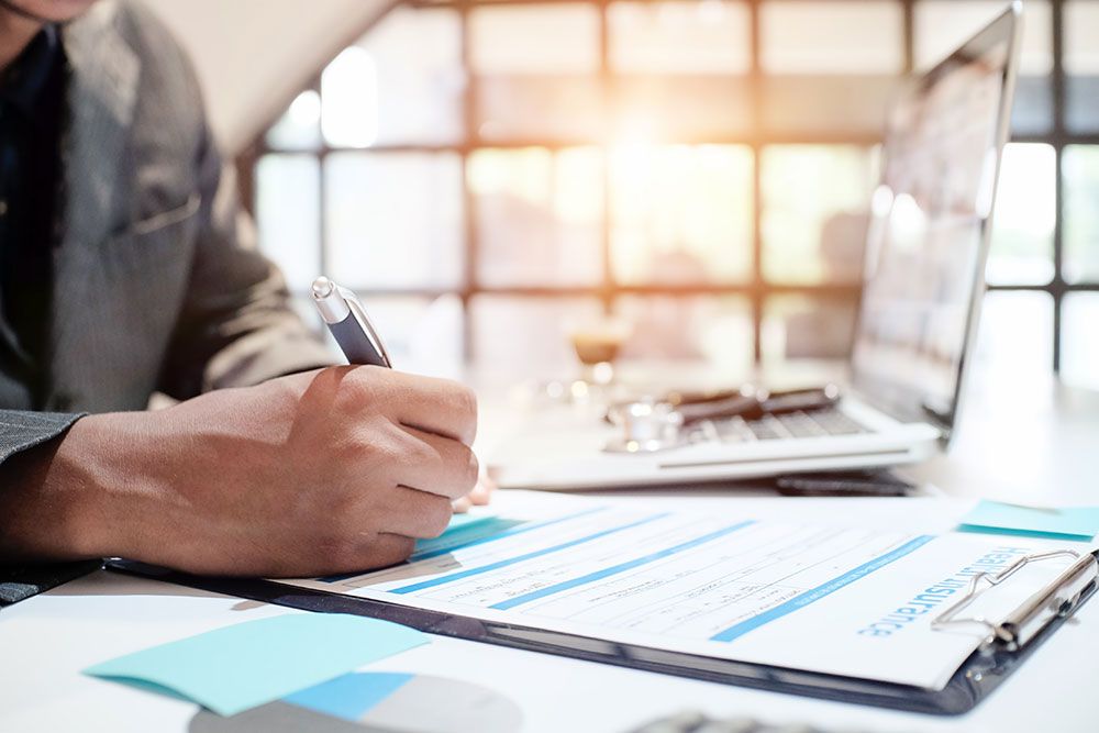 Person signing an insurance form at desk with laptop and clipboard, sunlight streaming through window.