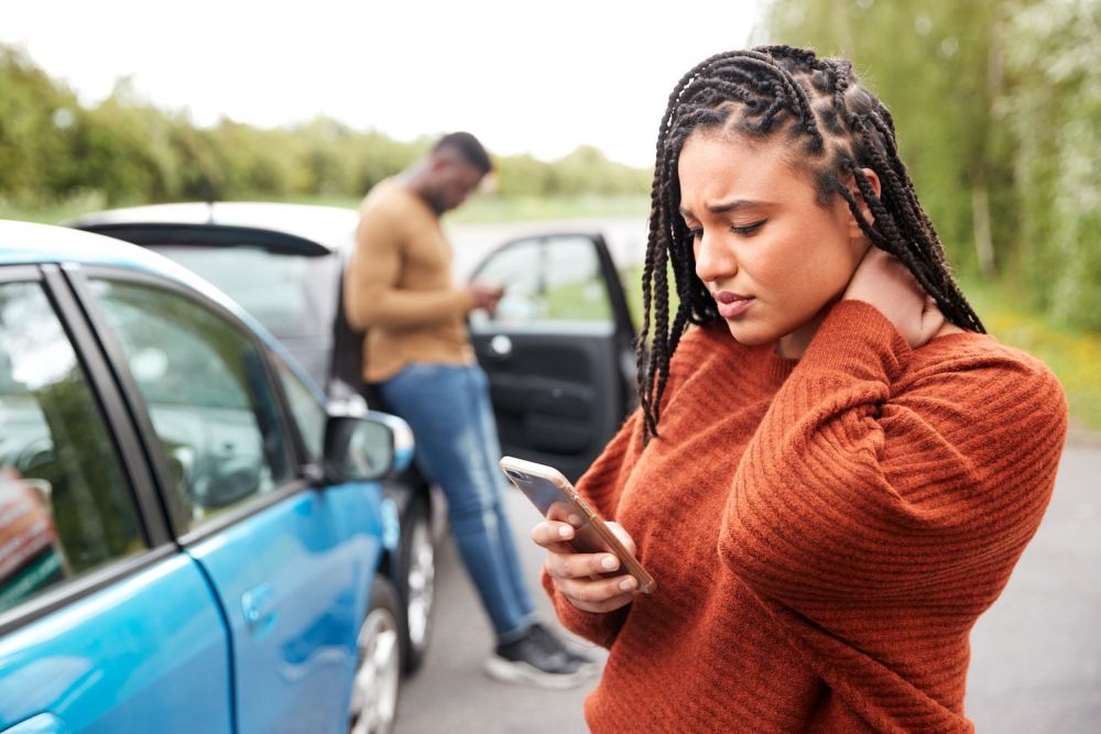 Woman on phone after car collision, rubbing her neck; man in background checking phone near car door.