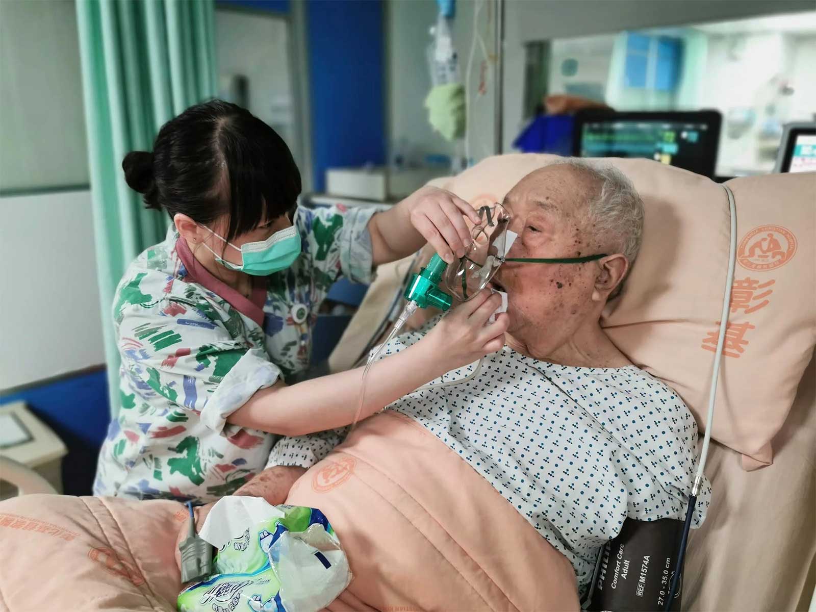 Female nurse adjusts elderly male patient’s oxygen mask while he’s in a hospital bed