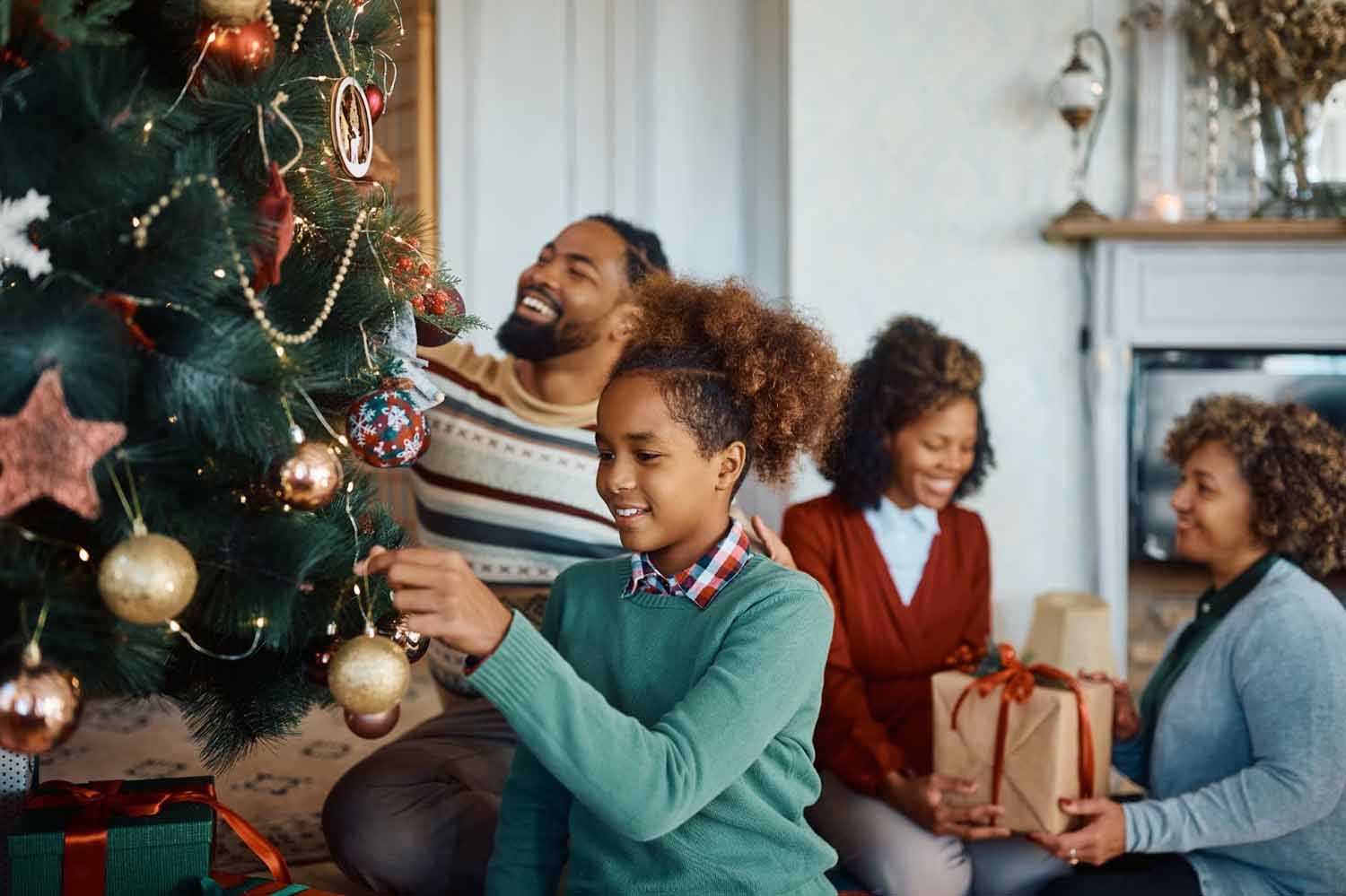 Happy family smiling and working together to decorate the Christmas tree and place wrapped gifts under the tree. The tree is placed in a safe place away from the fireplace.