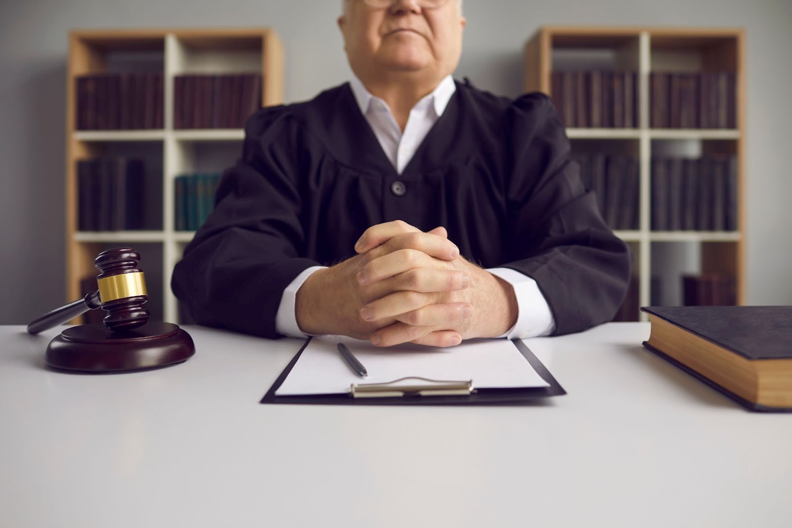 Older male judge with hands clasped at table with gavel and case papers in courtroom.