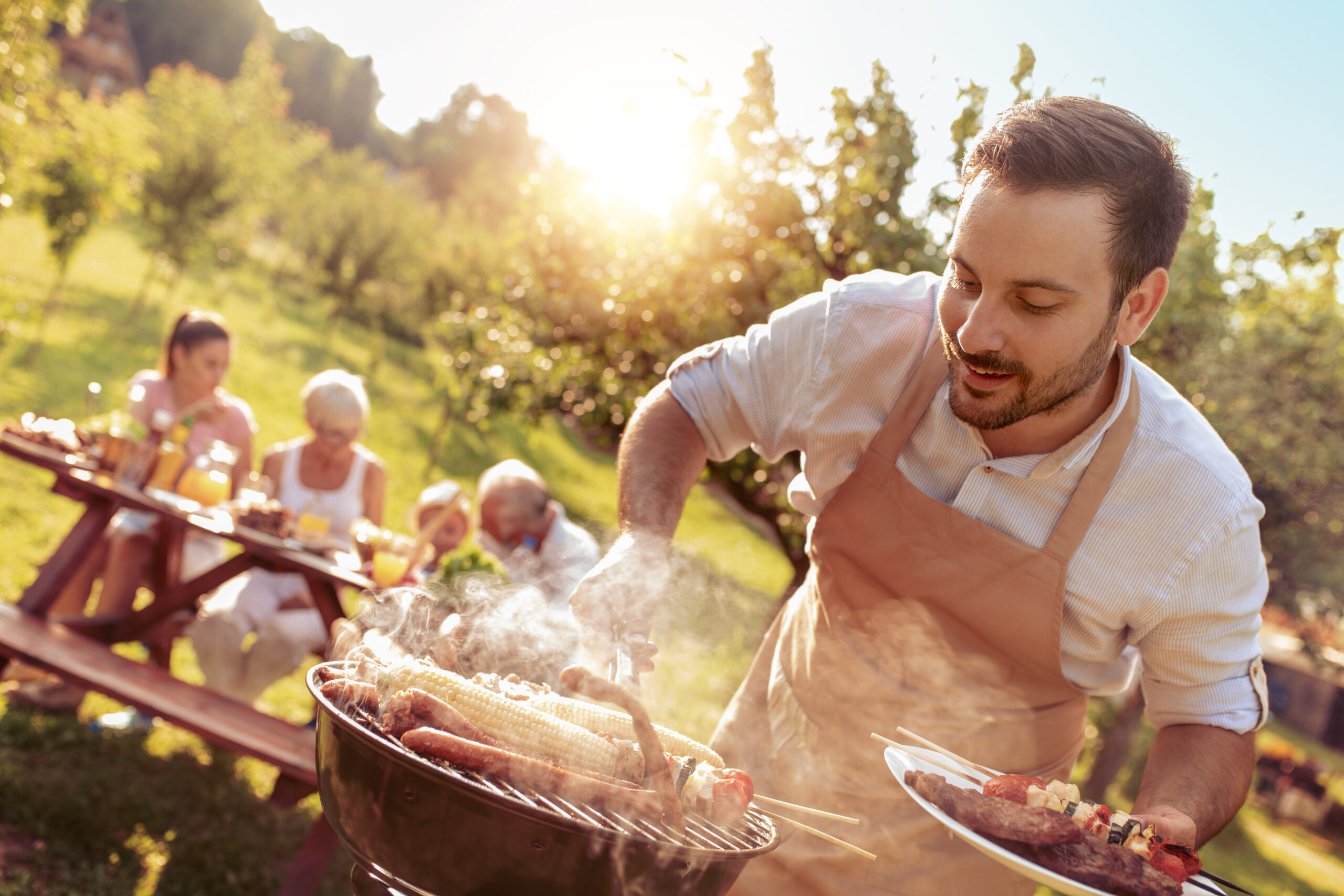 Man at the grill with family, blurred in the background at the table