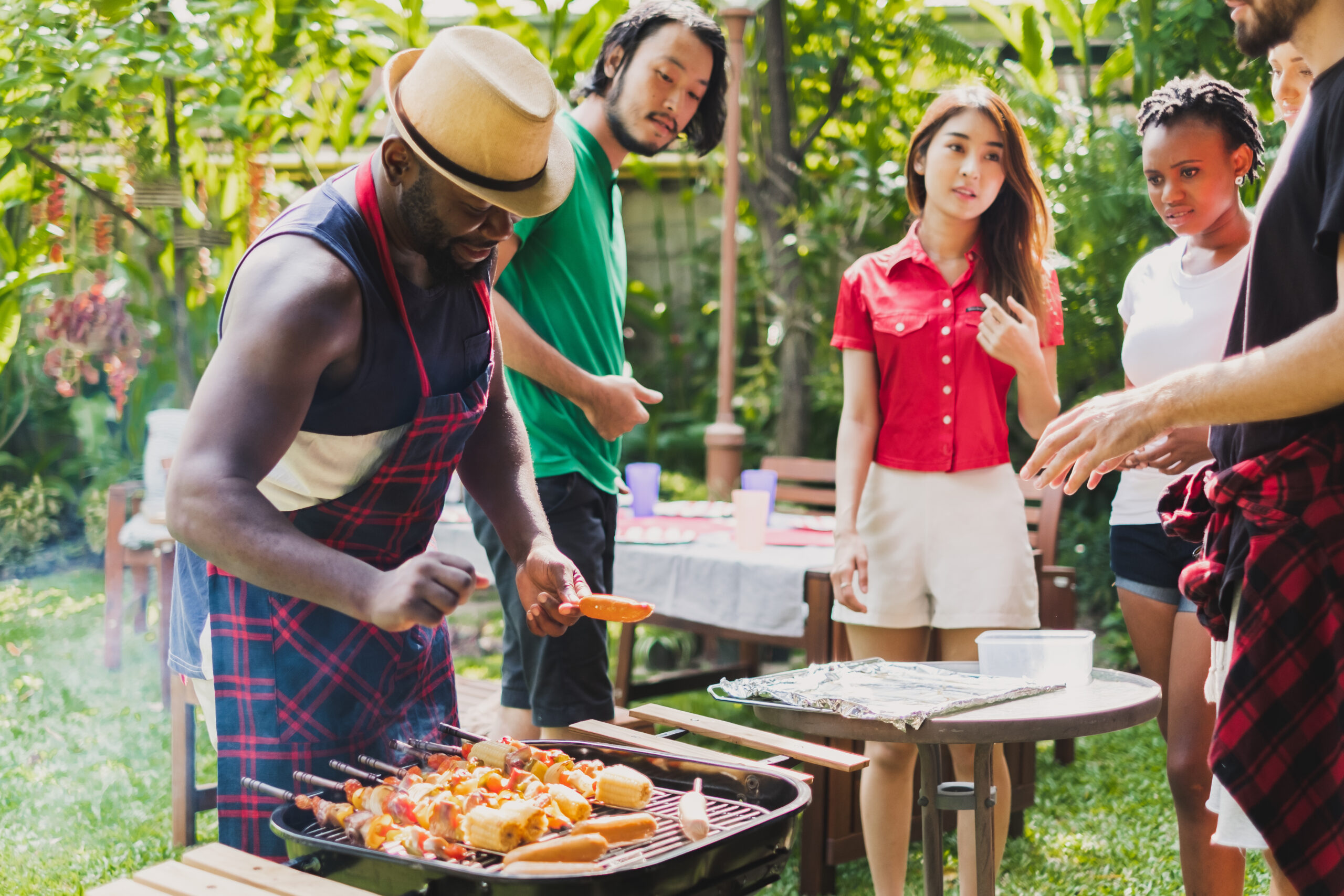 Man in hat cooking on the grill while his friends look on, ready to eat