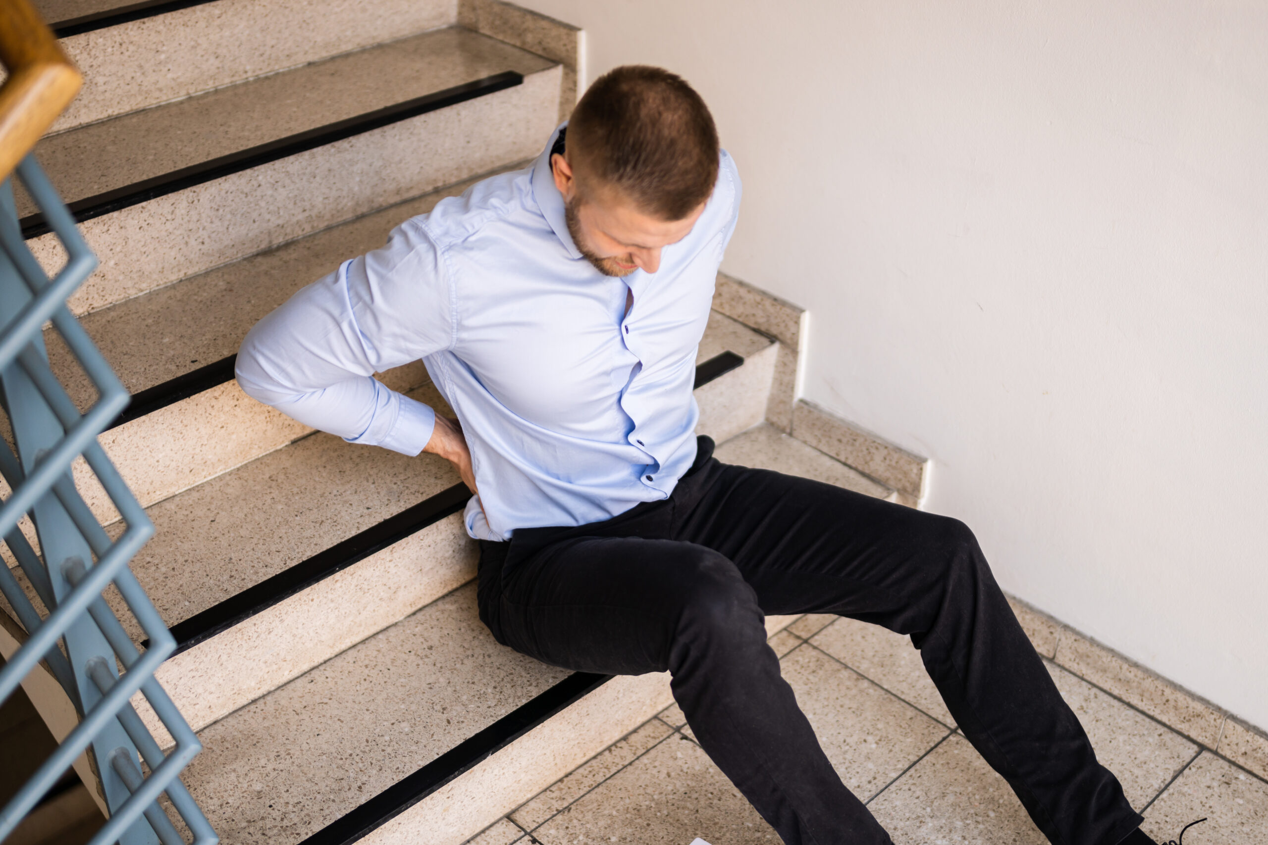 Man sitting at the bottom of the stairs, holding his back, wincing in pain - indicating he slipped and fell down the stairs