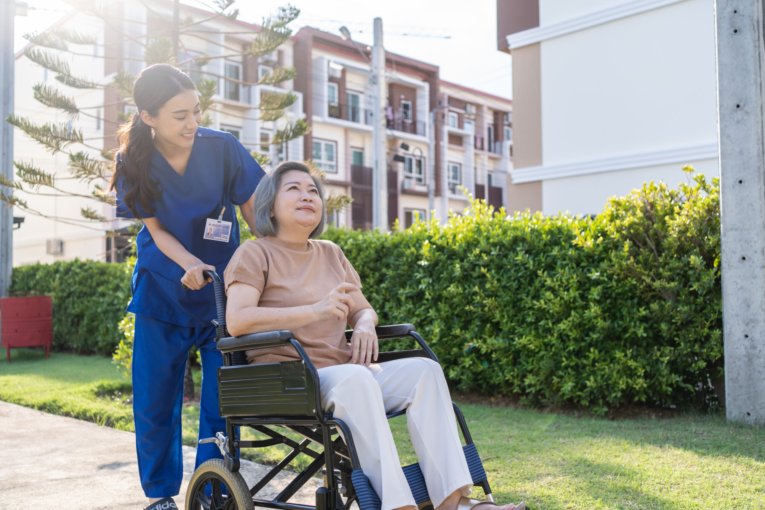 Female nursing assistant in blue scrubs pushes a disabled female patient in a wheelchair outside