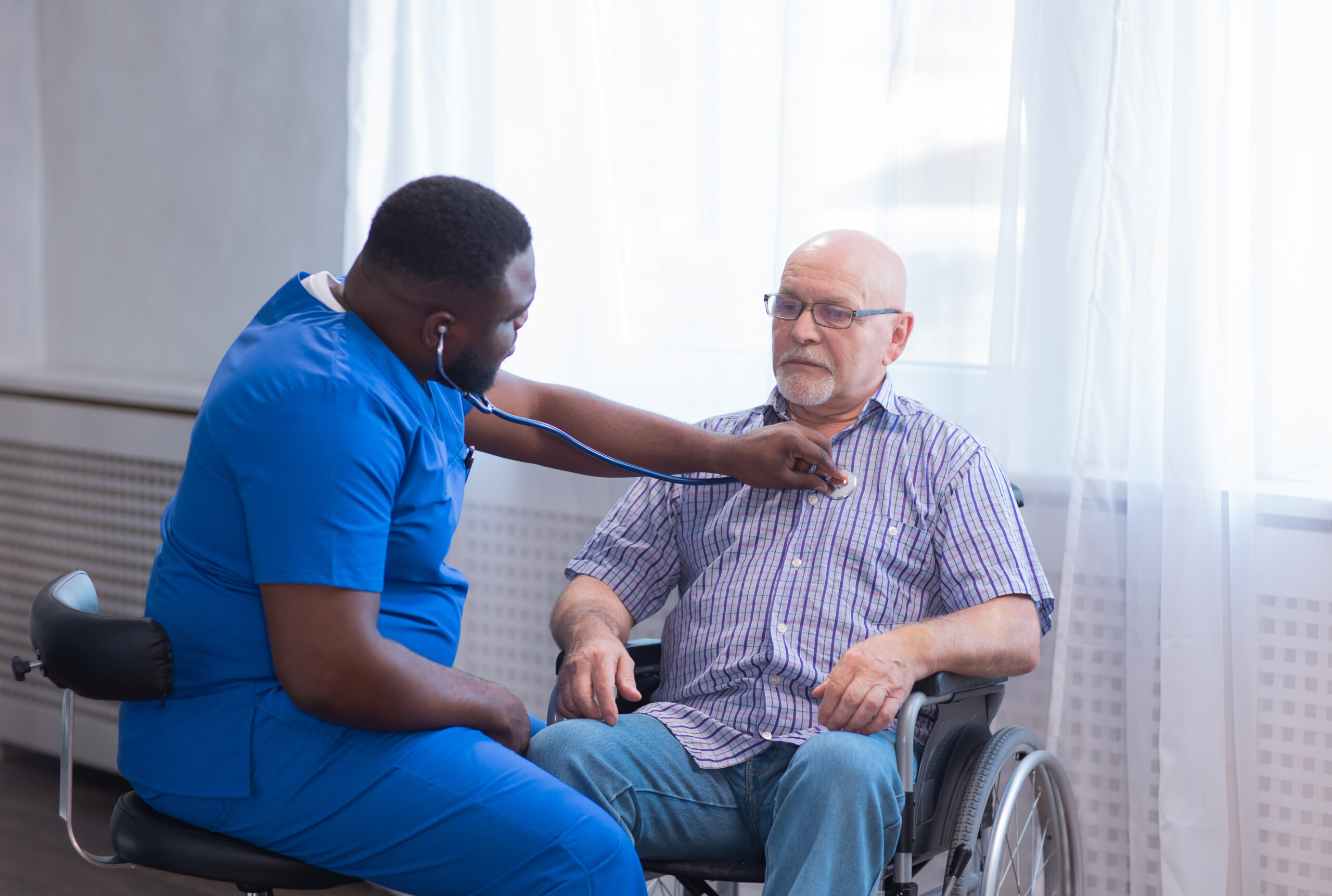 Male nursing assistant caregiver listening to the heart of an old man in a wheelchair with a stethoscope.