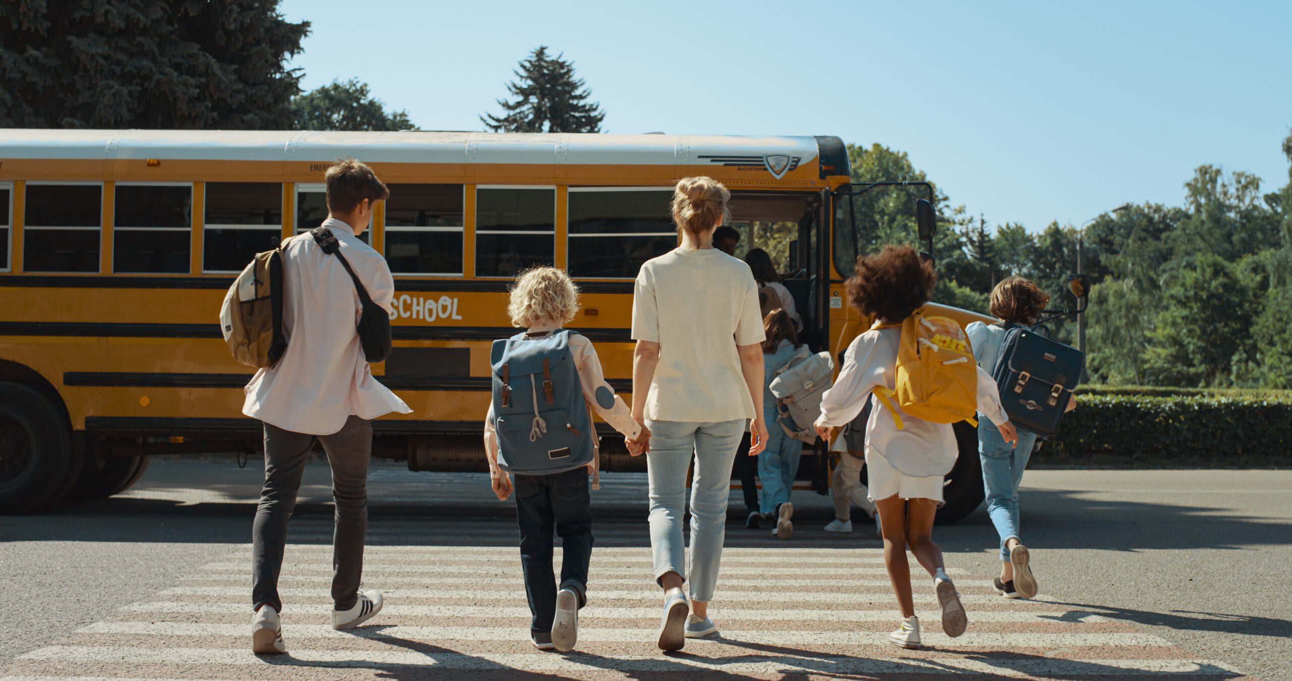 Mom holding son’s hand walking towards the school bus while eager children run by