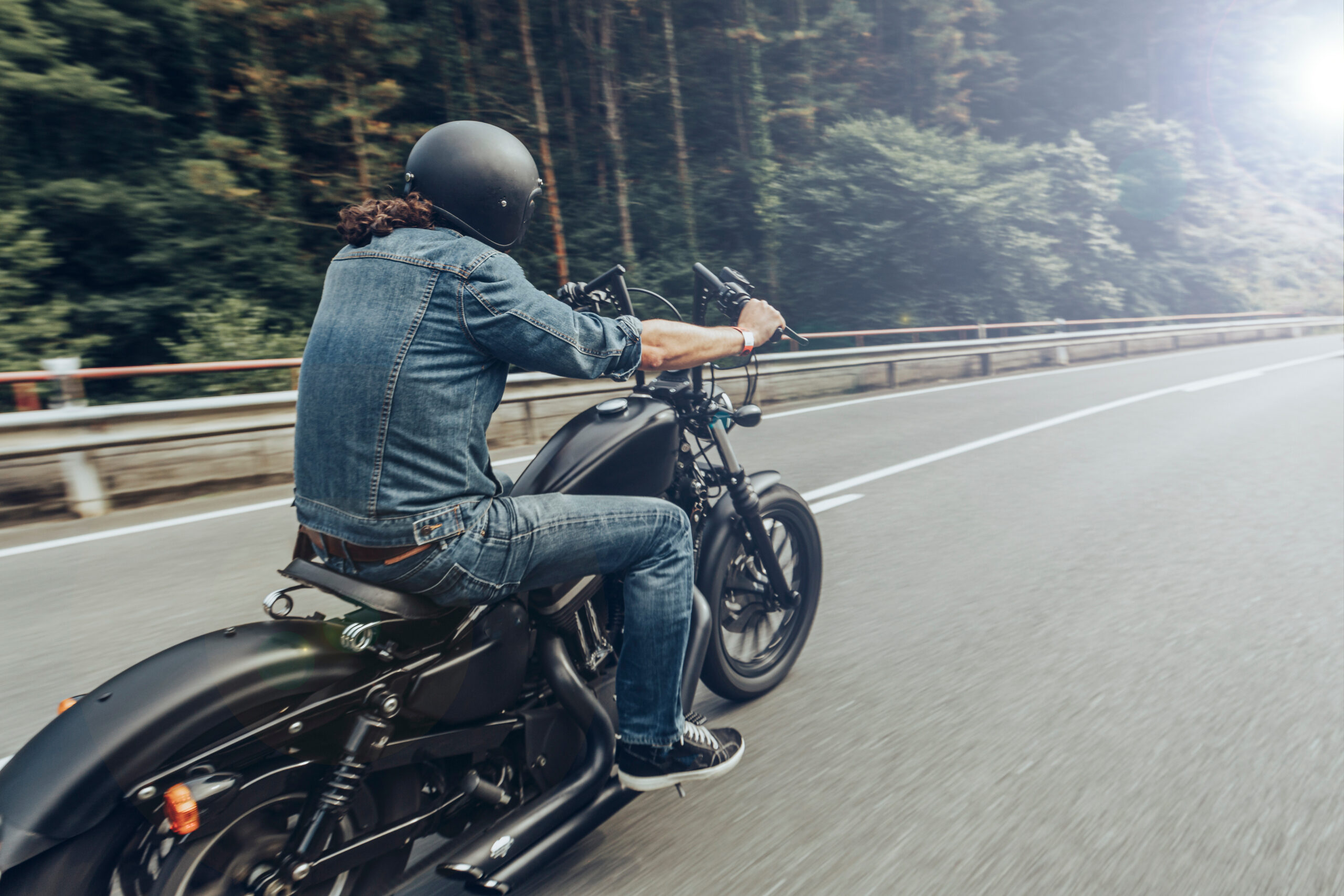 Motorcycle rider wearing a helmet, jean jacket and jeans, riding on a road near trees